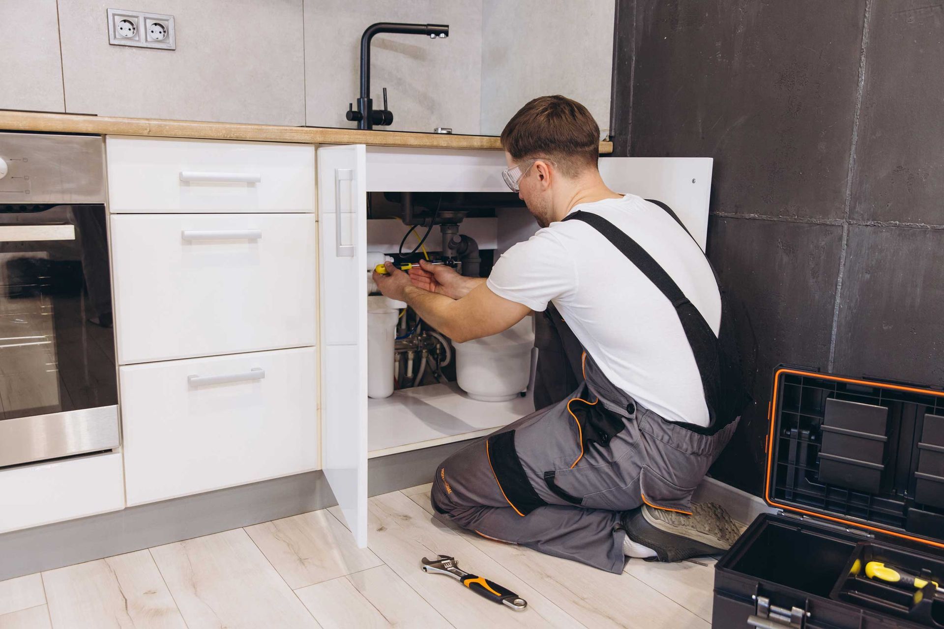 Licensed plumber repairing kitchen sink pipes under cabinet with toolbox and wrench nearby.