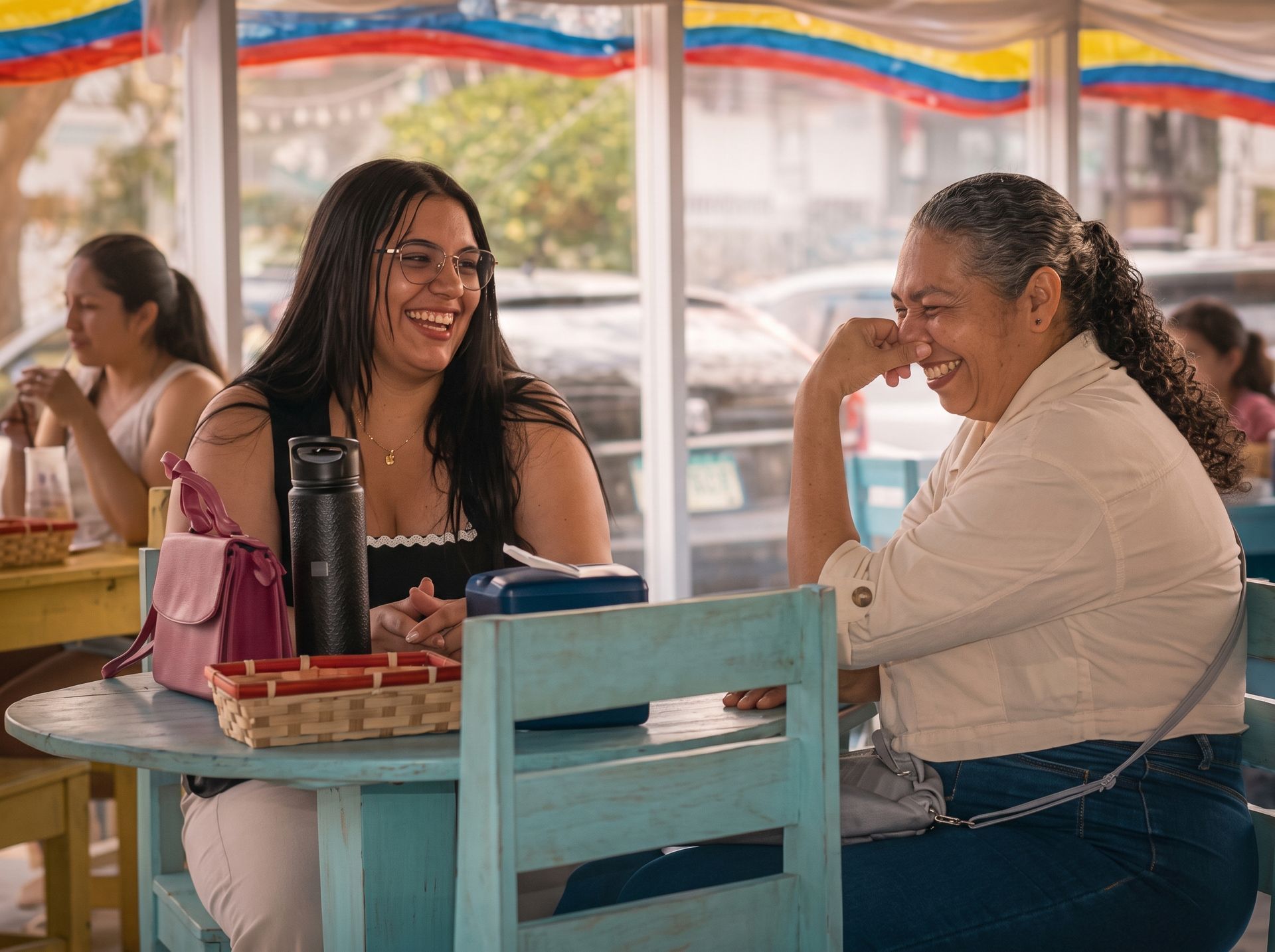 Two women smiling at a café table under a colorful awning