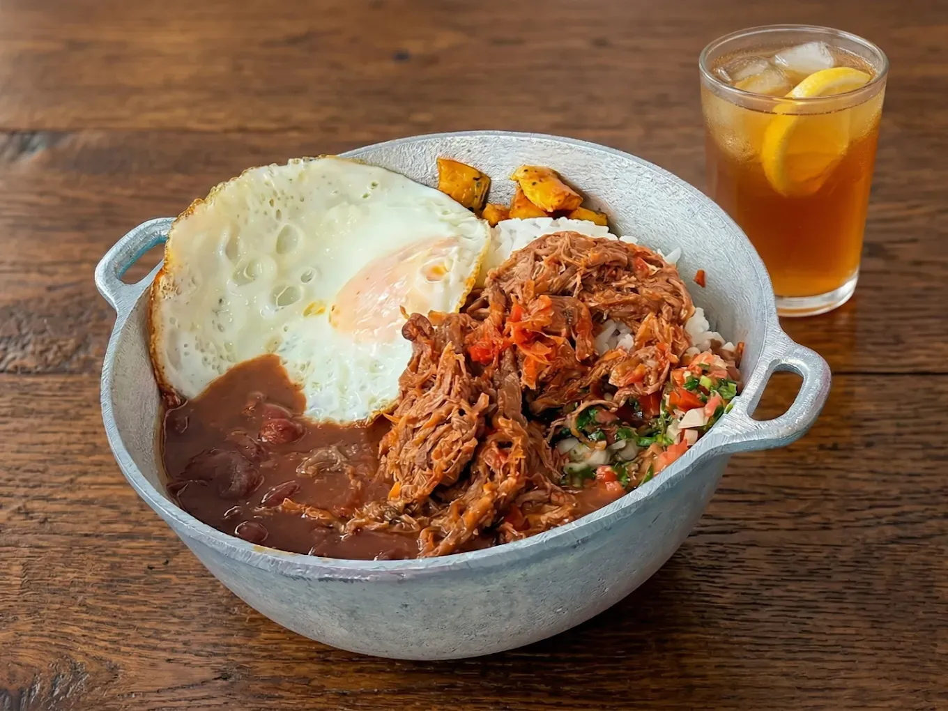 Bowl of rice, beans, shredded meat, fried egg, and plantains with a glass of iced tea on a wooden table
