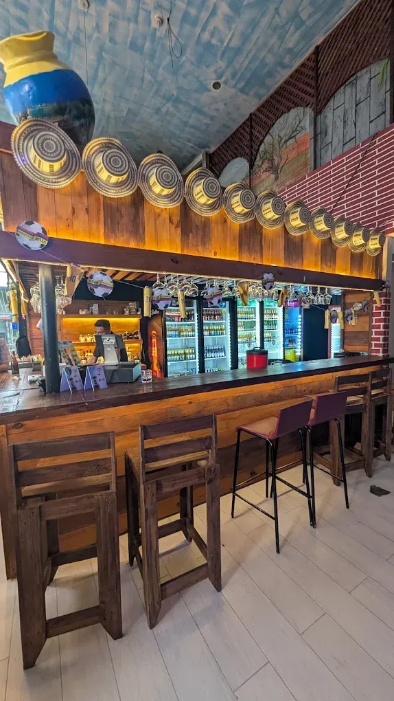 Empty bar with wooden stools, lit shelves, hanging glasses, and patterned ceiling tiles.