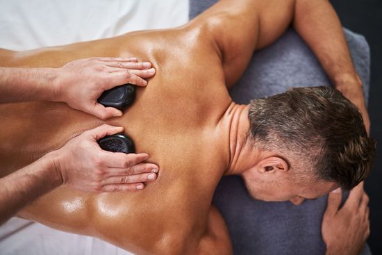 Hot stone massage on a person’s back with hands arranging black stones and white flowers nearby