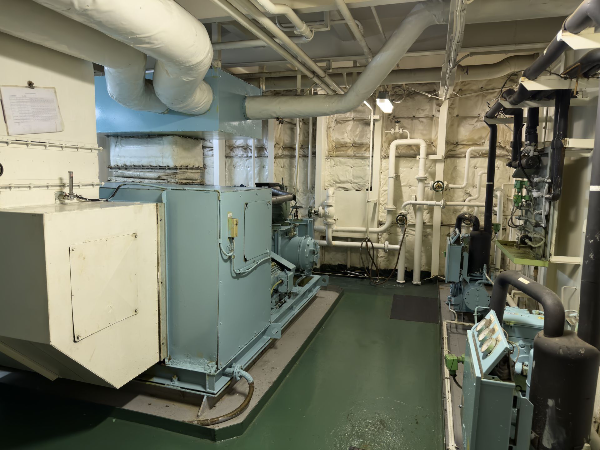Industrial machinery in a cramped boiler room with white pipes and green flooring