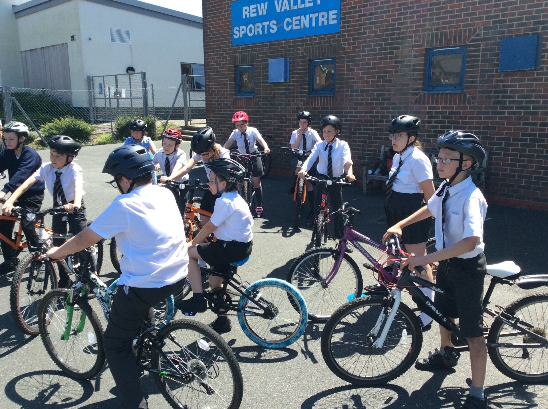 Students in uniform with helmets on bikes outside New Valley Sports Centre. Sunny day.