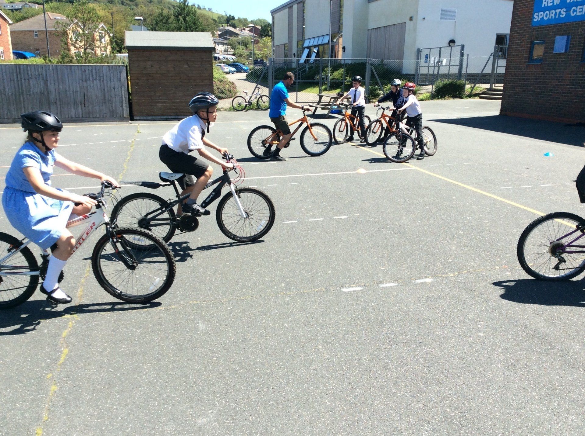 Children riding bikes in a schoolyard, wearing helmets. A blue-shirted adult supervises. Sunny day.