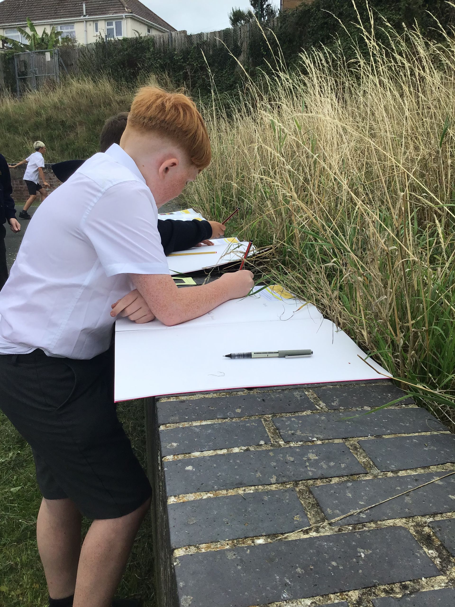 Person in white shirt writing on paper at an outdoor table beside tall grass