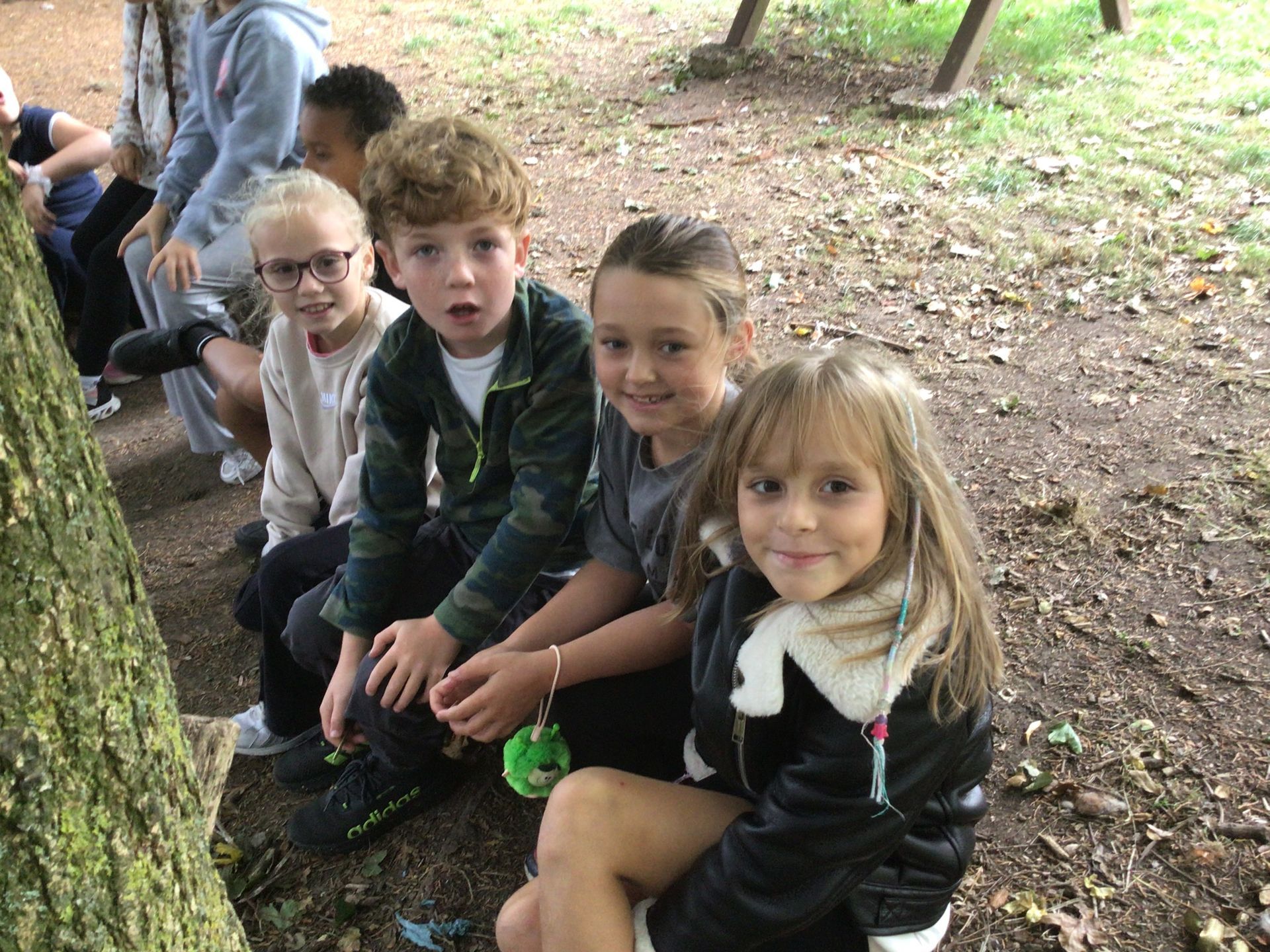 Four children sitting on a bench outdoors, facing the camera, with trees and grass around them.
