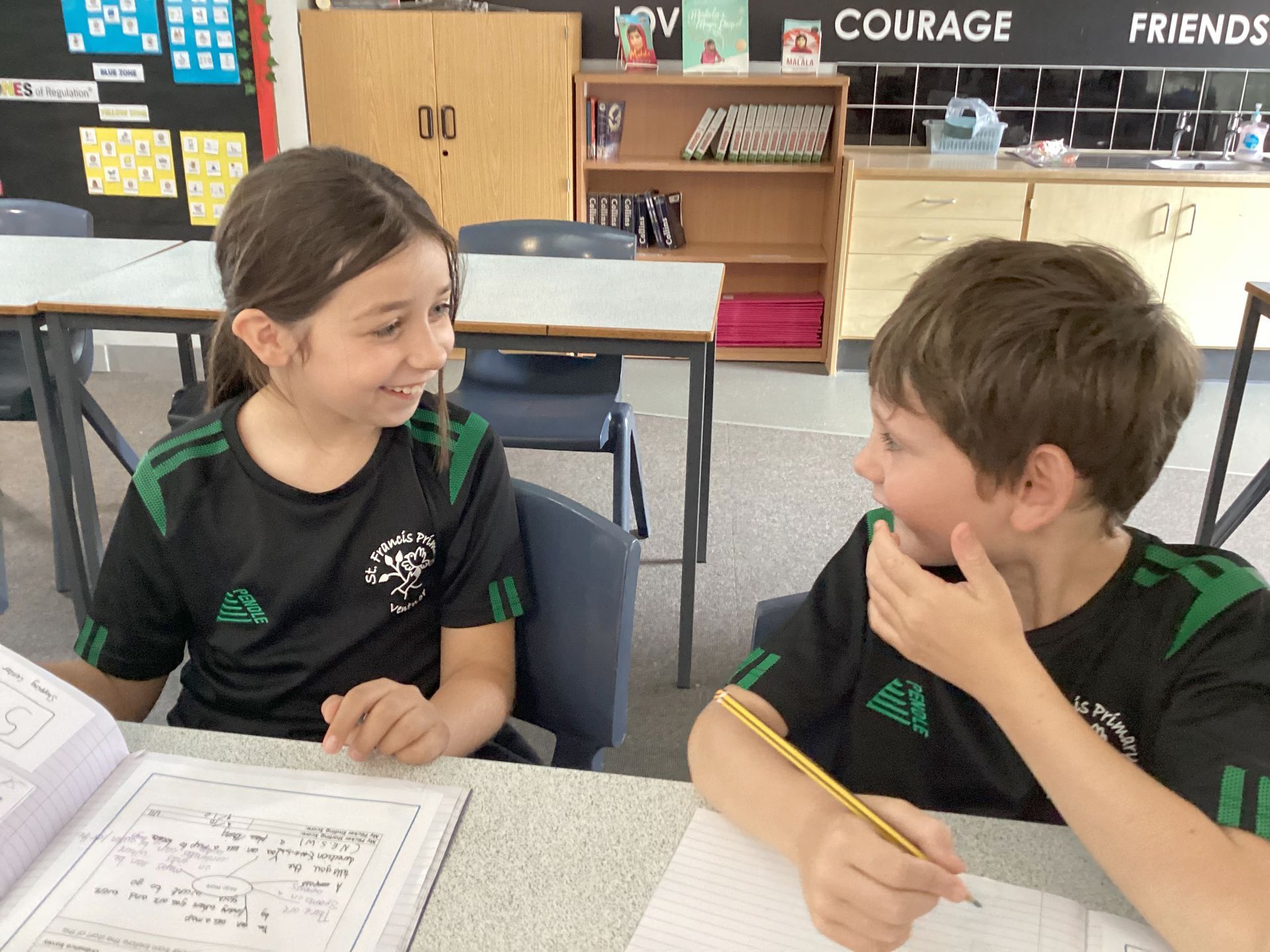 Two students smiling at each other across a classroom table, with papers and pencils in front of them.