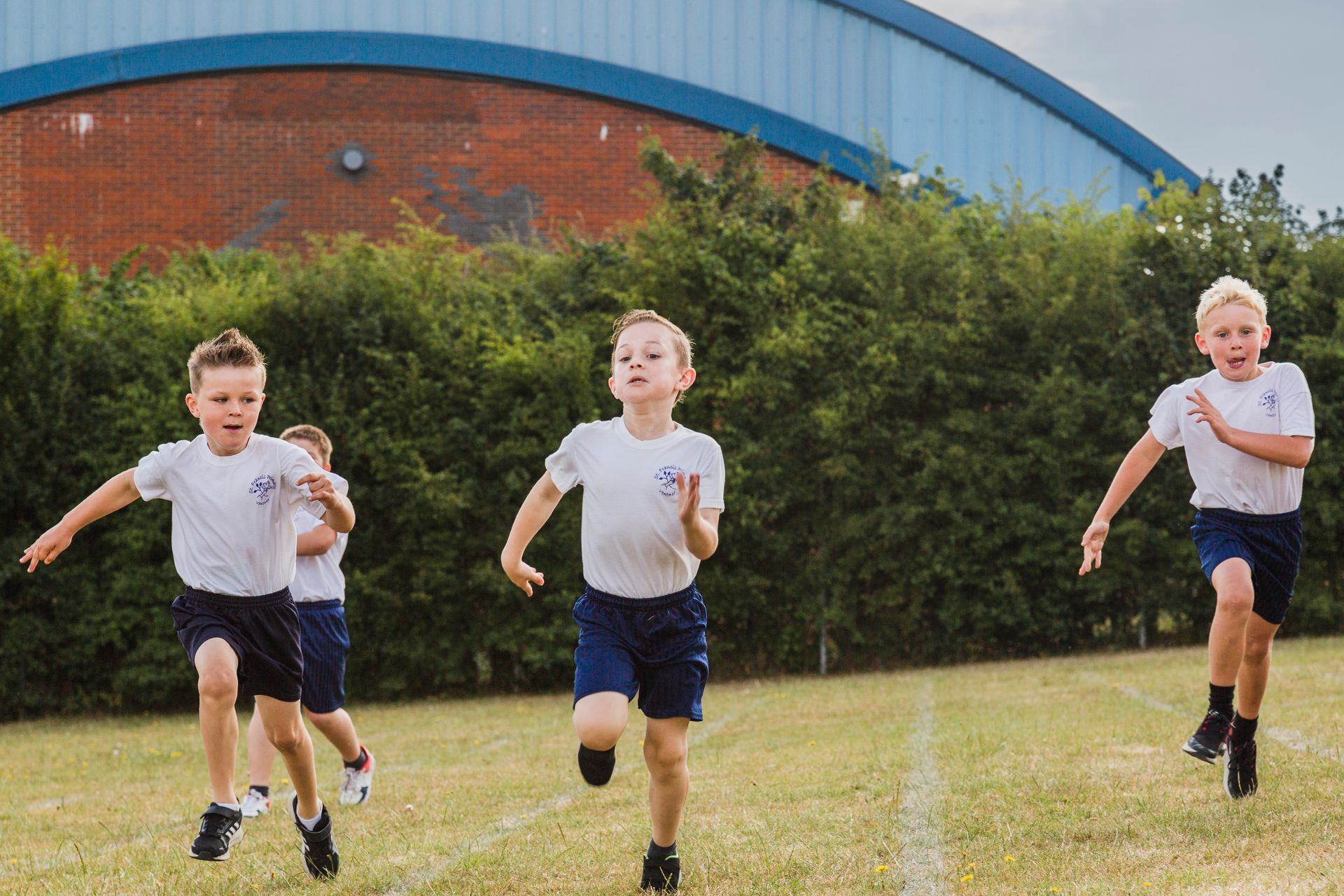 Boys in school uniforms running on a grass track, near a building.
