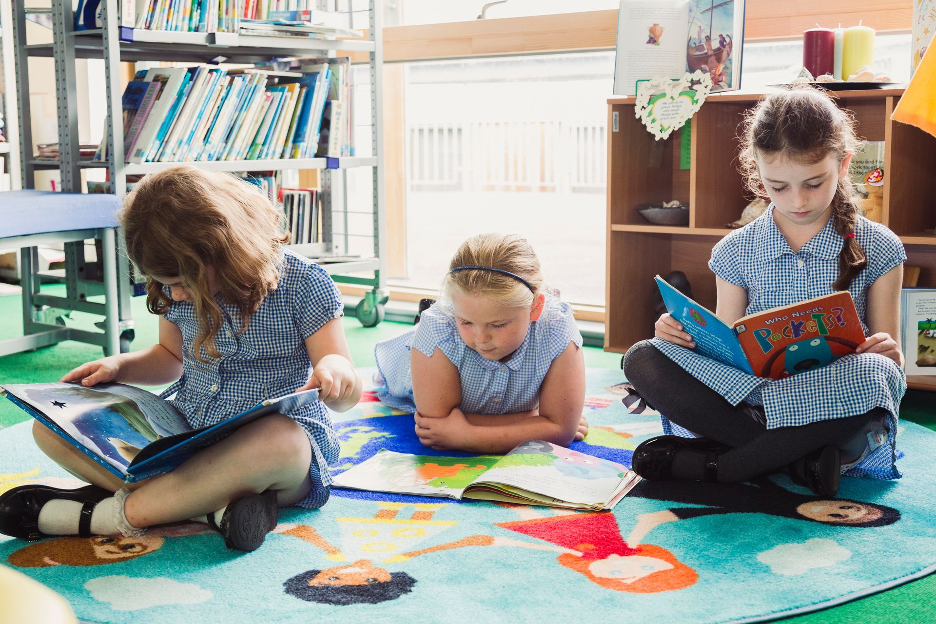 Three young girls reading books on a colorful rug in a library.