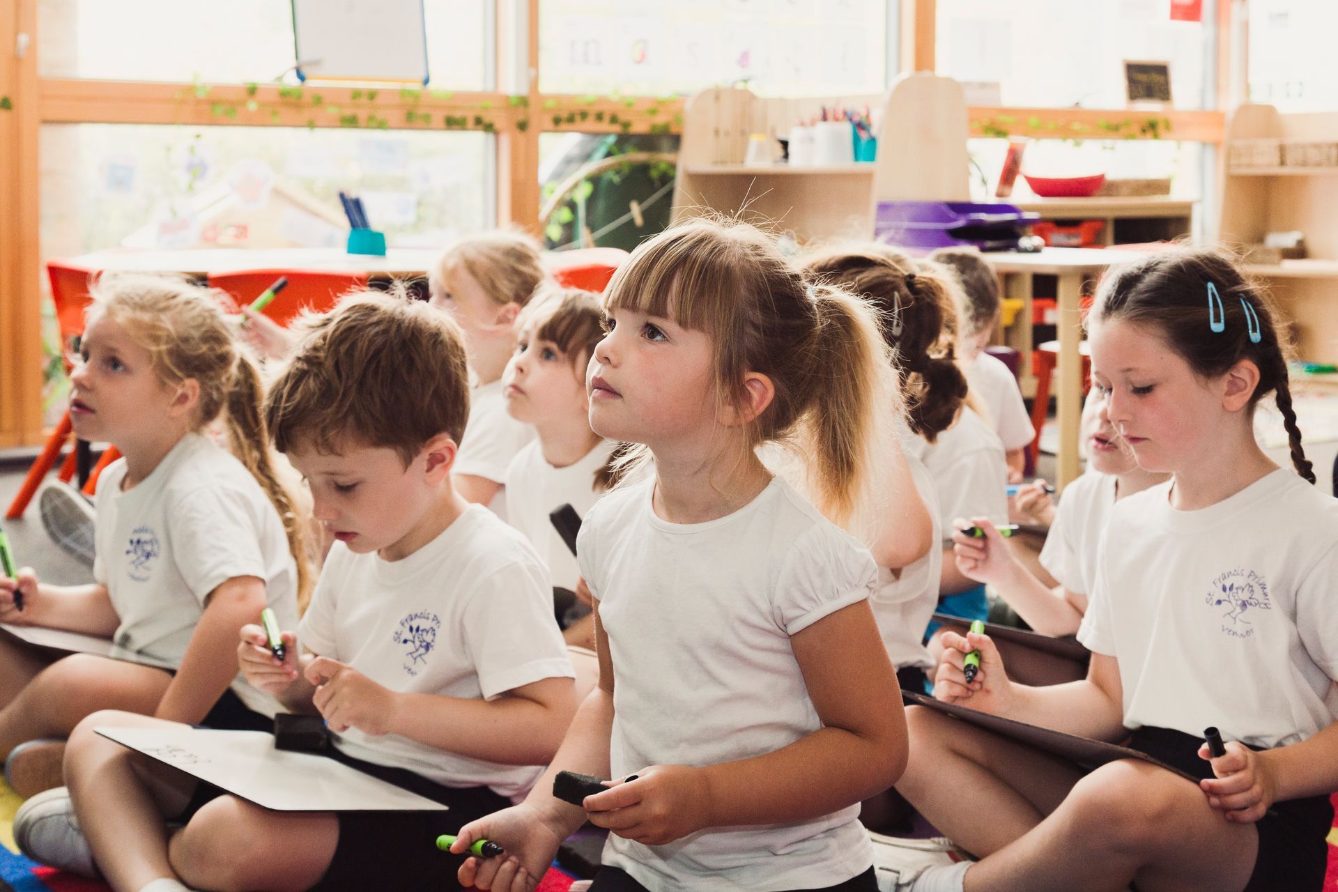 Children in white shirts sit on the floor, looking up, holding markers and drawing boards in a classroom.