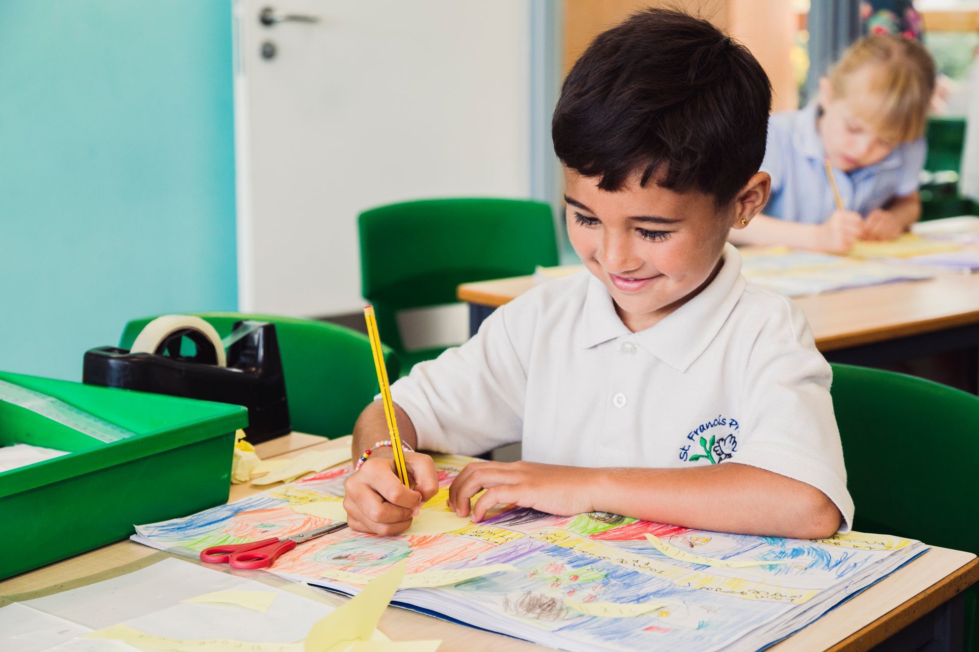 Boy in white shirt drawing at desk in a classroom, smiling.
