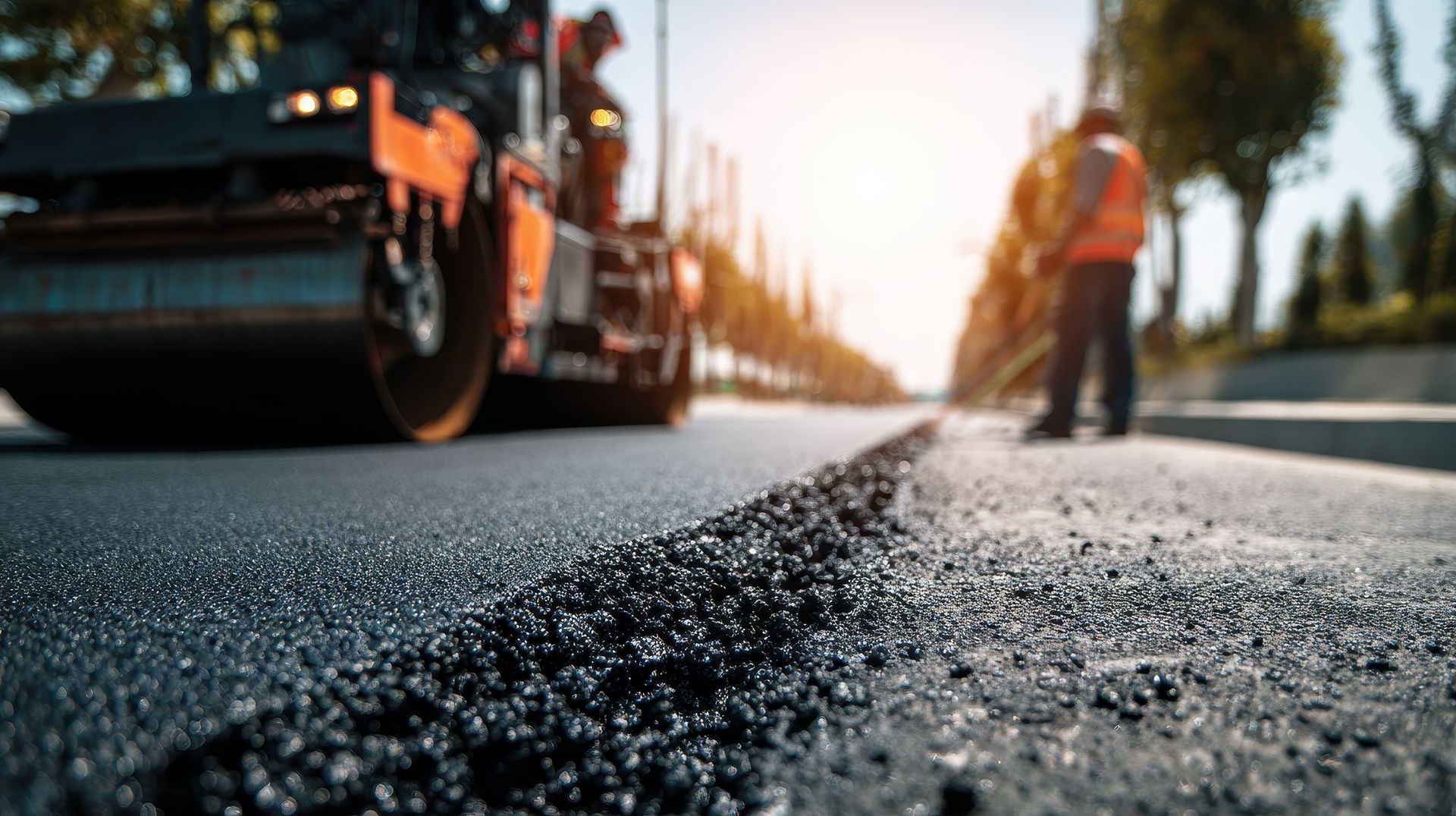 Road paving in progress; asphalt roller compacting freshly laid blacktop, worker in vest observing.