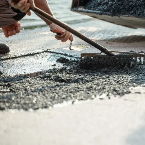 Road paving in progress; asphalt roller compacting freshly laid blacktop, worker in vest observing.