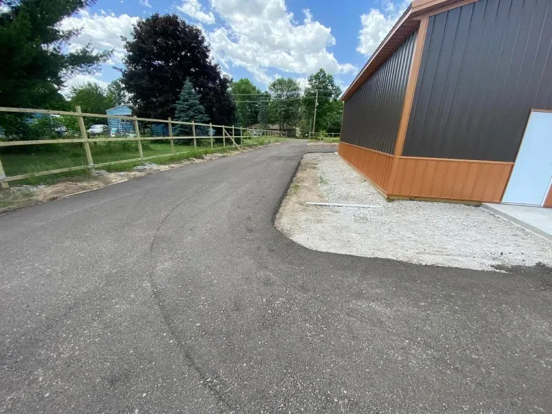 Gravel path beside a brown metal building, with a wooden fence and trees under a cloudy sky.