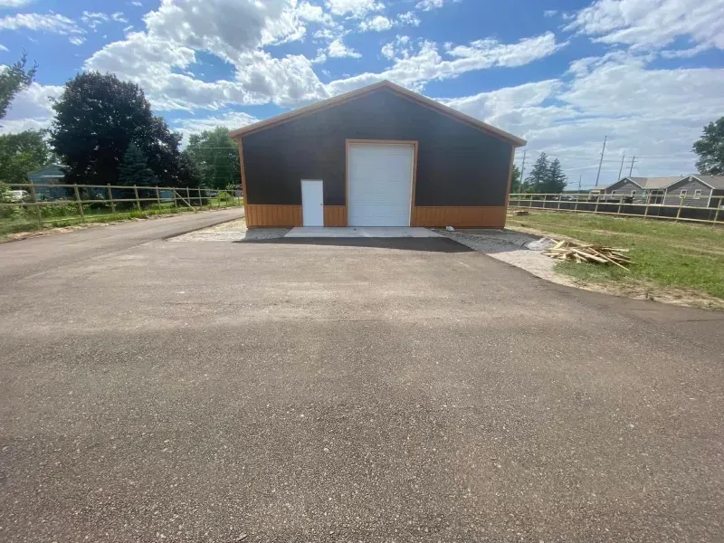 Small brown building with a white garage door at the end of a paved lot under a cloudy sky
