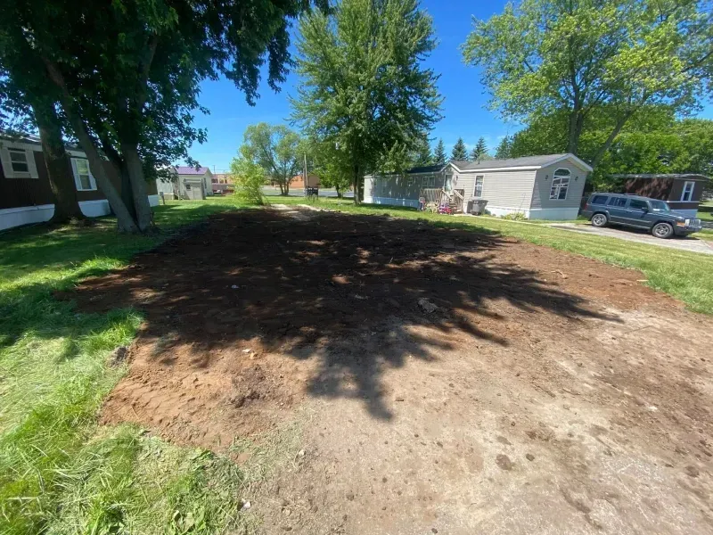 Dirt lot with a large shaded area, surrounded by trees, mobile homes, and parked vehicles on a sunny day