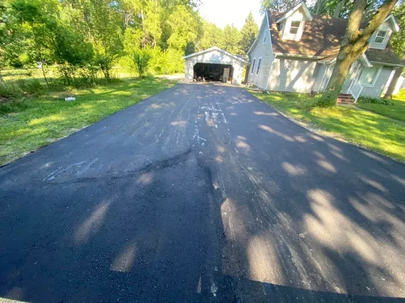 Freshly paved driveway leading to a garage beside a white house and green trees in bright sunlight