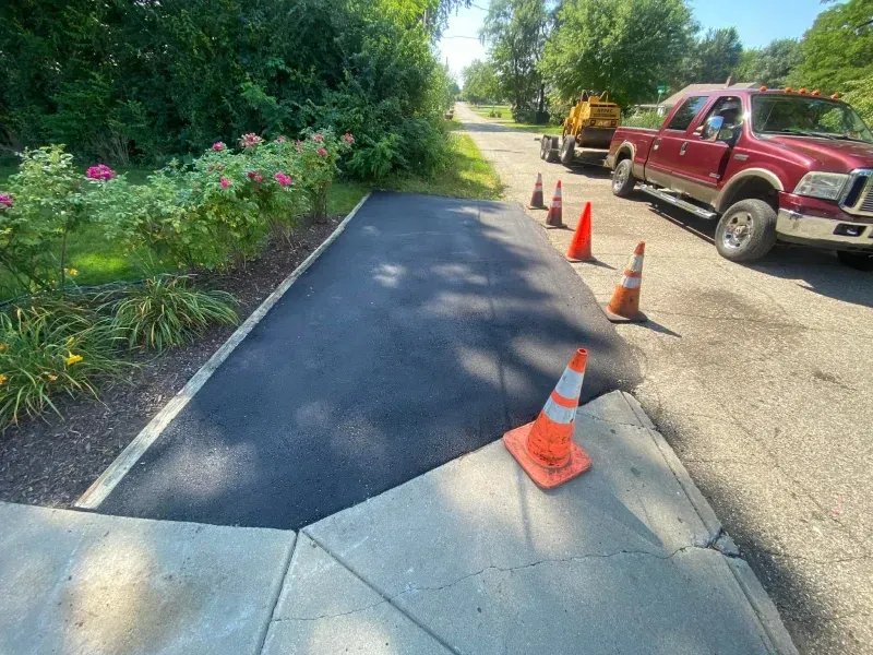 Freshly paved driveway beside a garden, with orange cones and a red truck parked on the gravel shoulder