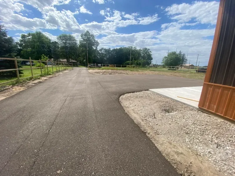 Paved driveway beside a wooden building, with gravel edge, fence, trees, and cloudy blue sky.