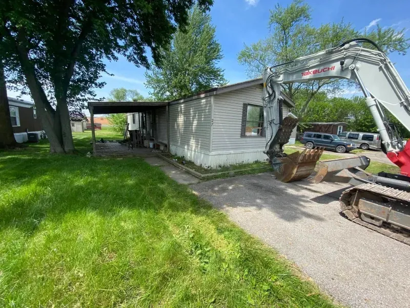 Small white house with a covered carport, grassy yard, and an excavator beside the driveway.
