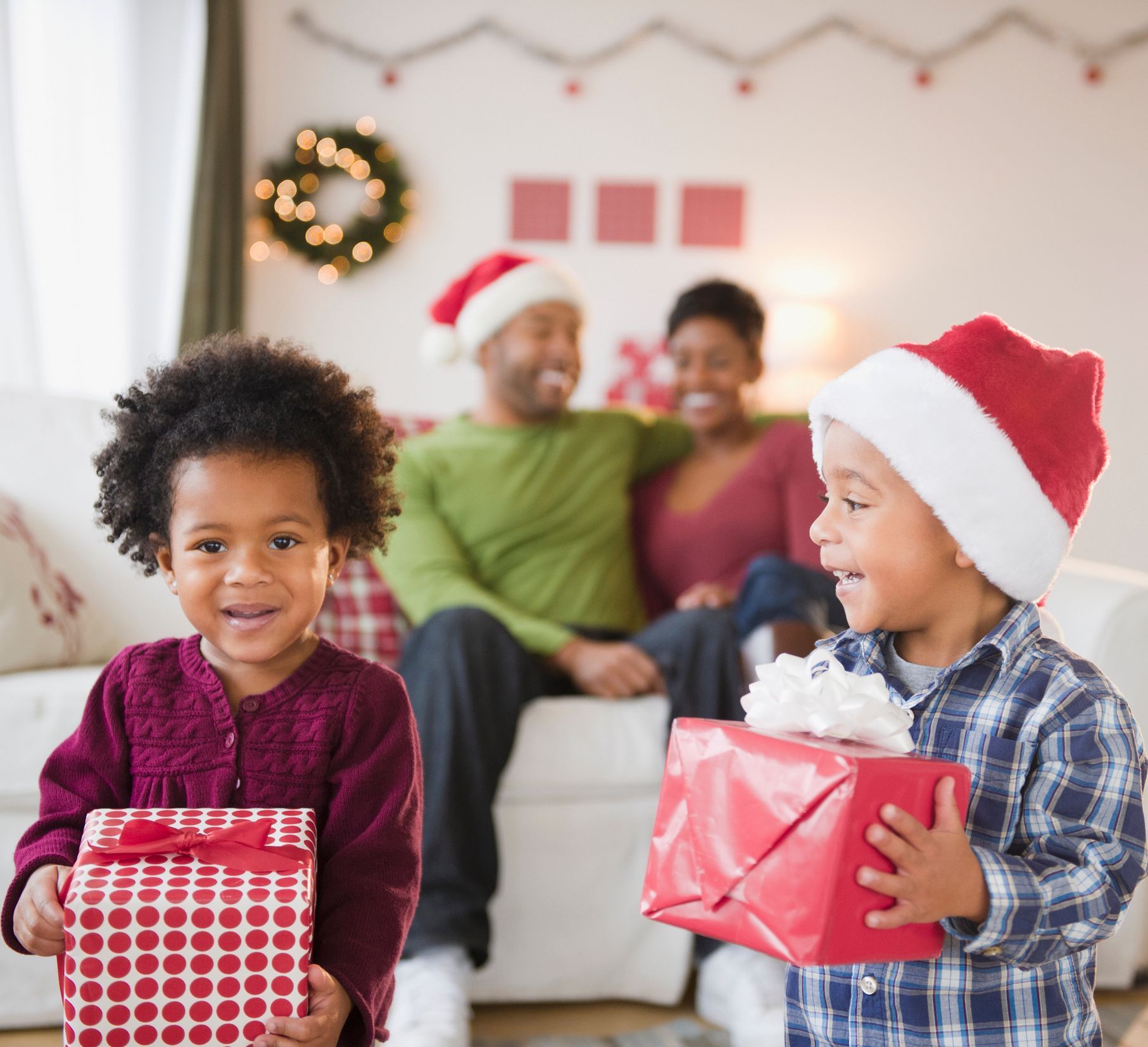 Children holding gifts smile at camera, family in background on Christmas.