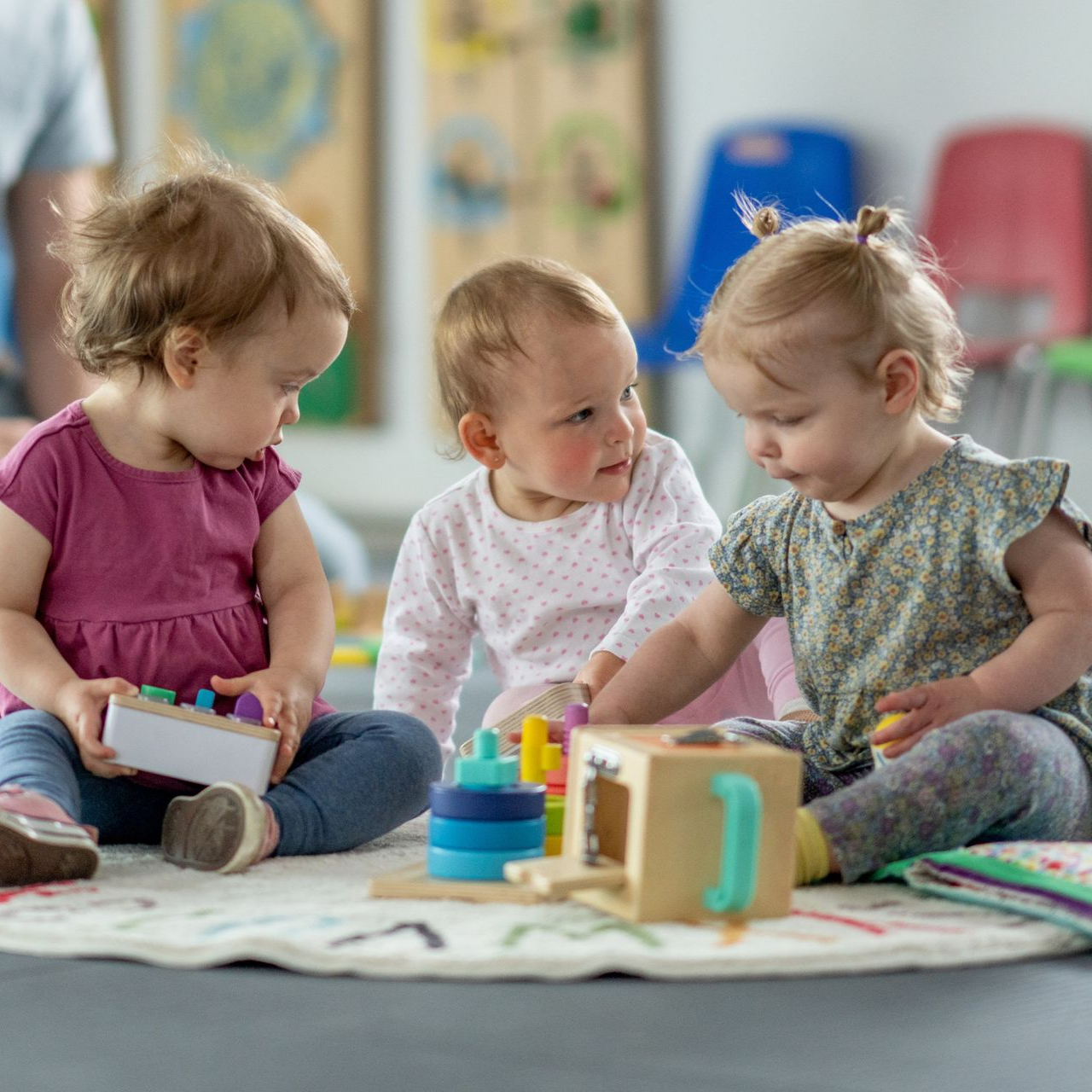 Three toddlers playing with wooden toys on a colorful mat indoors.