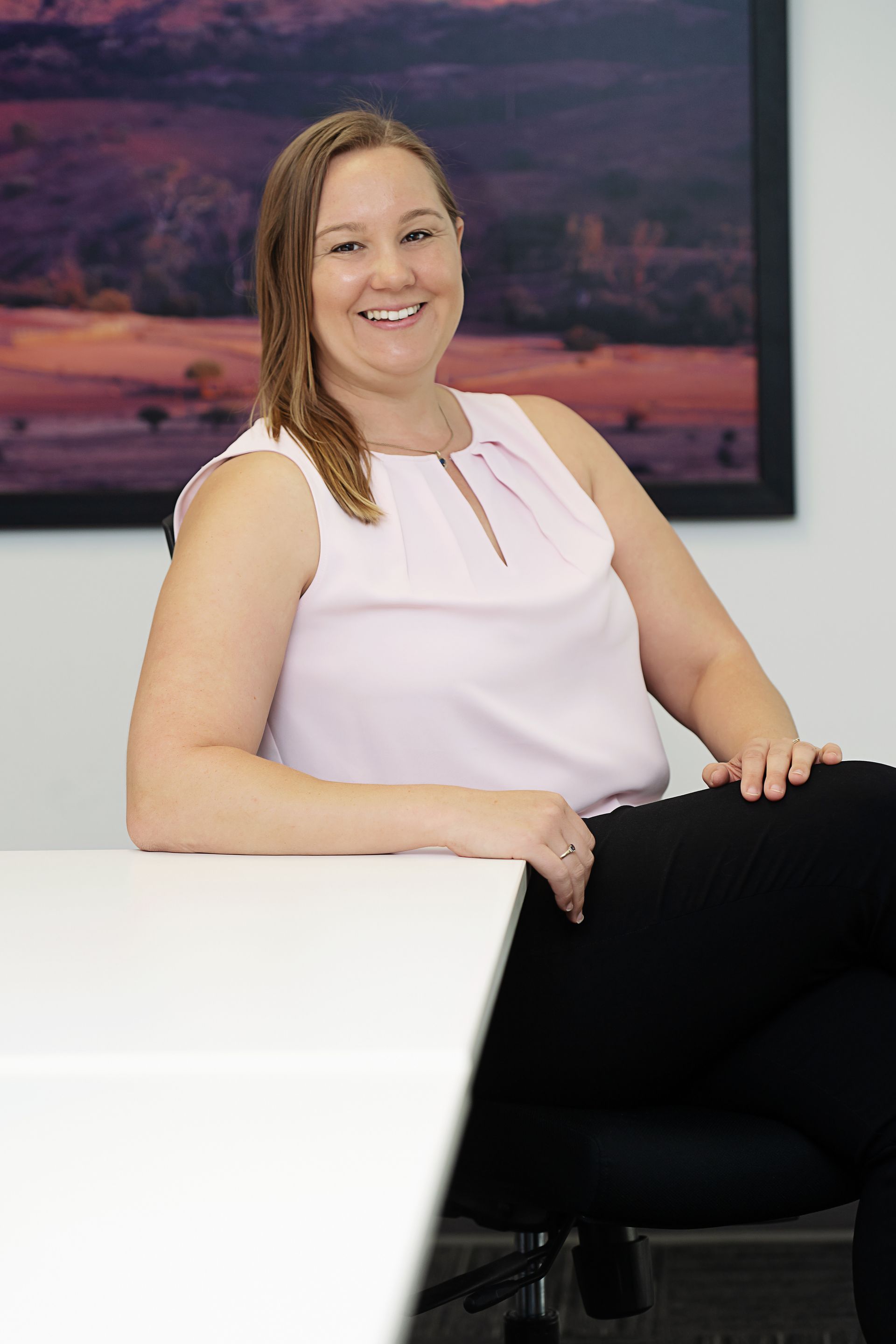 Smiling woman in a light pink top seated beside a white table in an office.