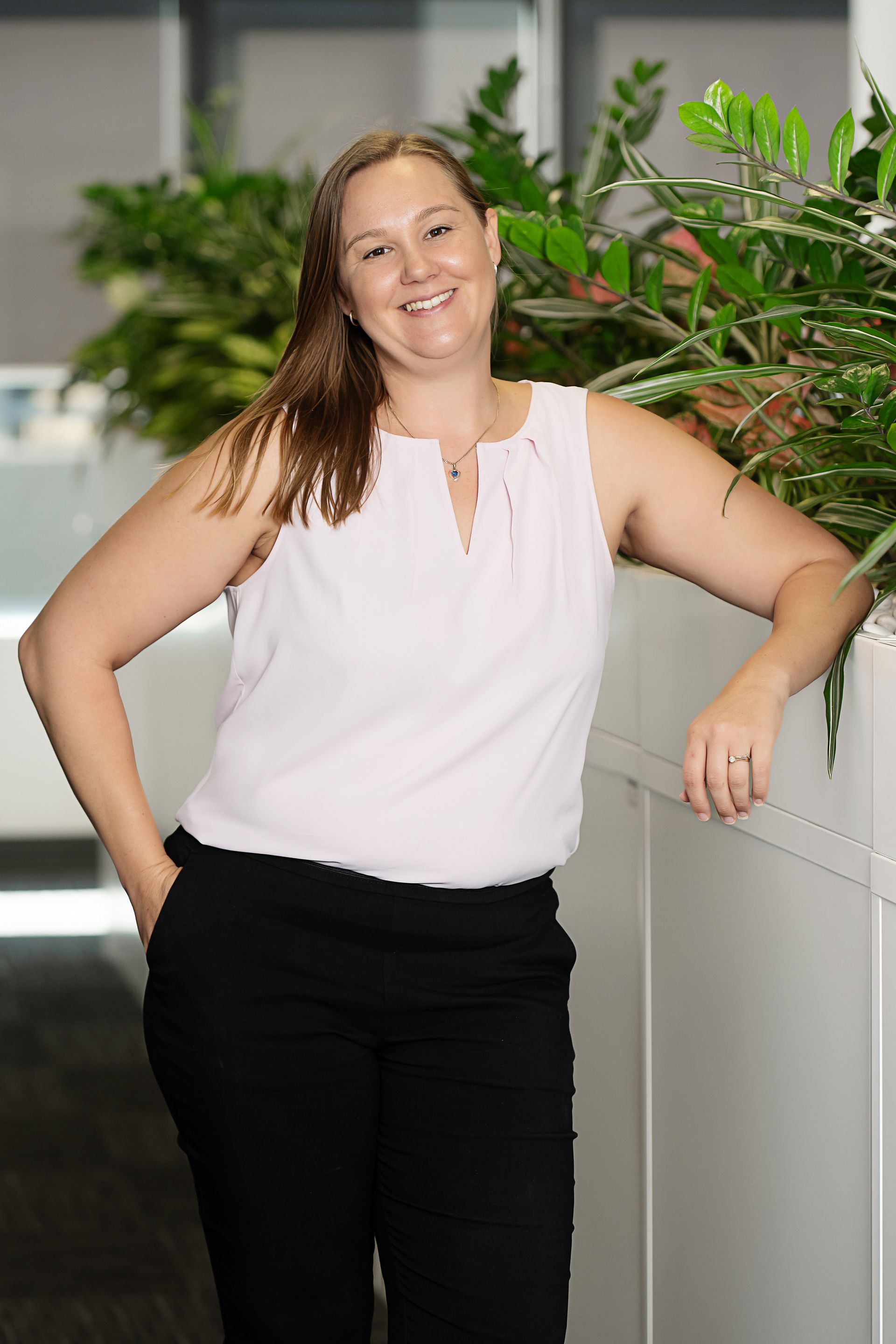 Woman in a white sleeveless top and black pants leaning against a railing with plants in an office setting