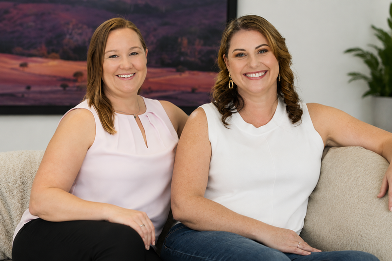 Two smiling women sitting on a couch in a living room, wearing light tops.