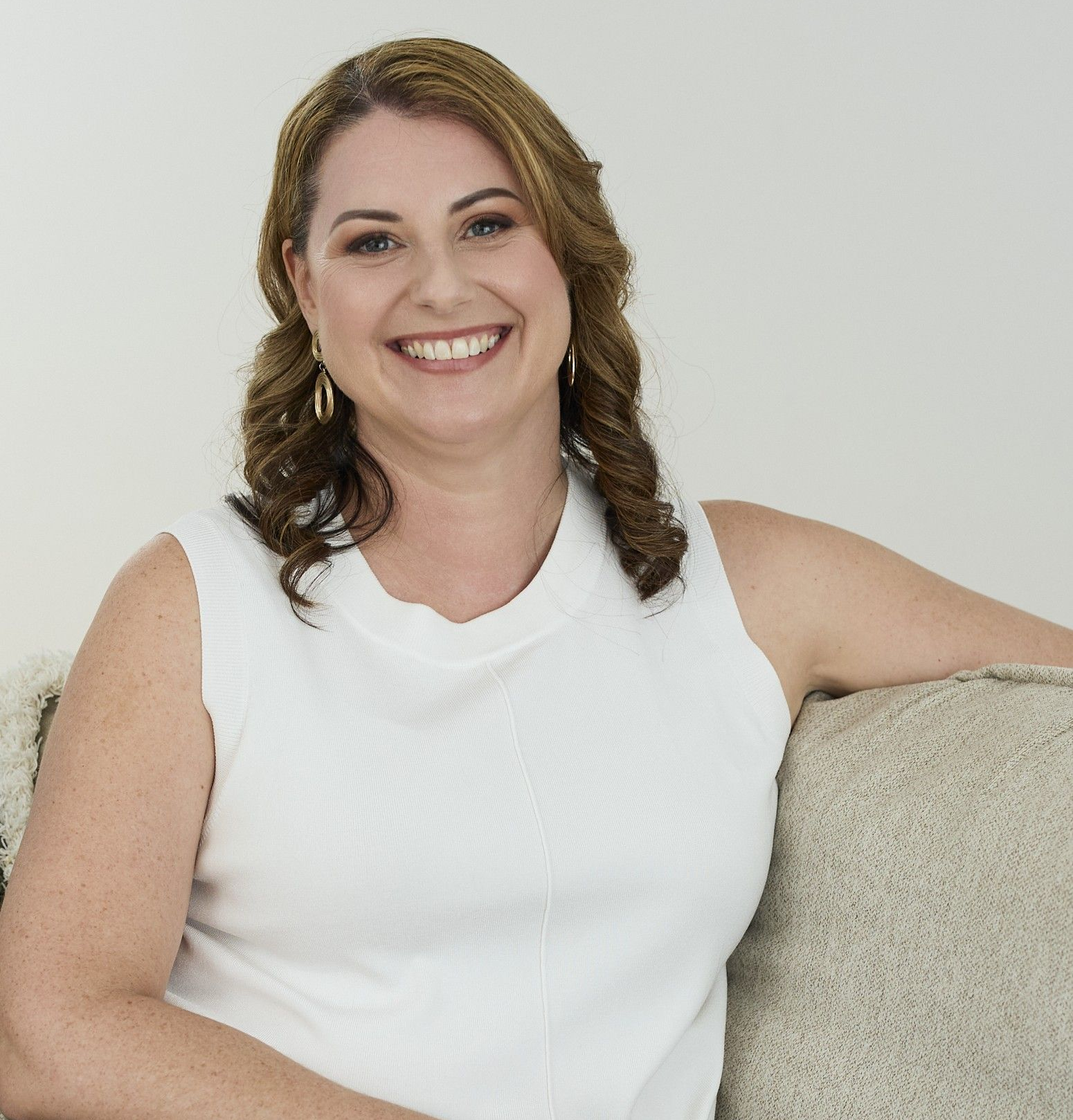 Smiling woman in a white sleeveless top sitting on a couch against a light background