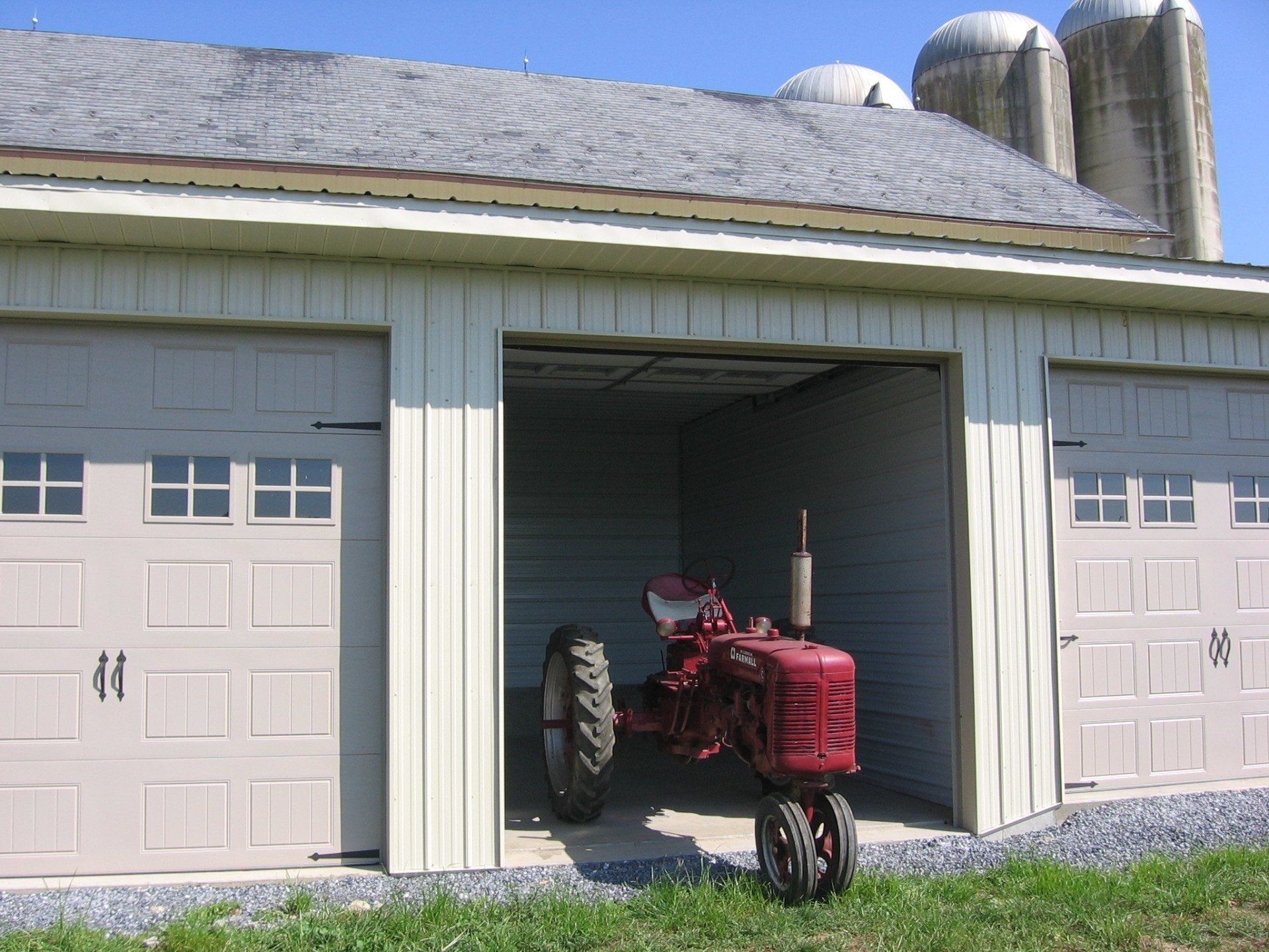 Red tractor in an open garage with two white doors on a farm building.
