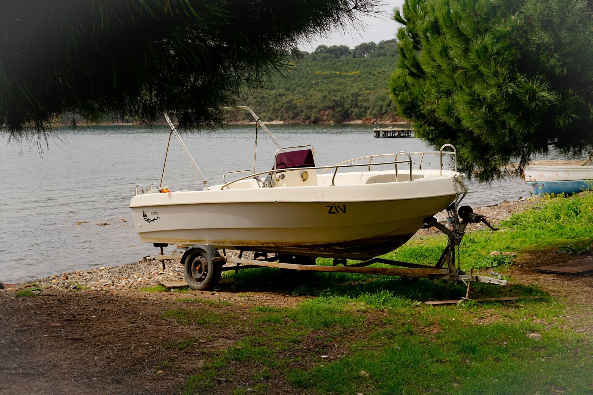 Small white motorboat on a trailer by a lakeshore, beside a tree and grassy bank.