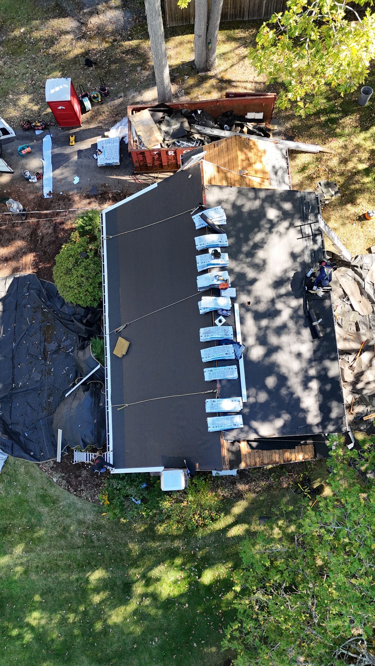 An aerial view of a house roof undergoing repairs, with materials staged along the ridge and debris on the ground below.