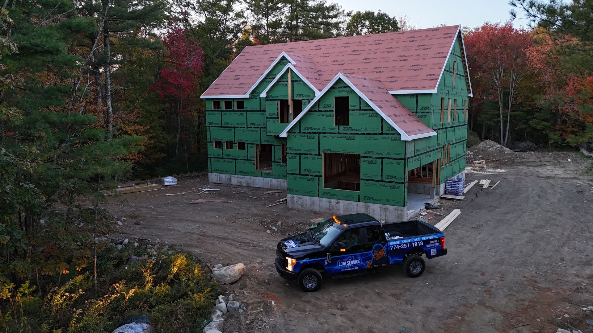 A dark pickup truck parked in front of a new two-story house under construction, surrounded by autumn trees.