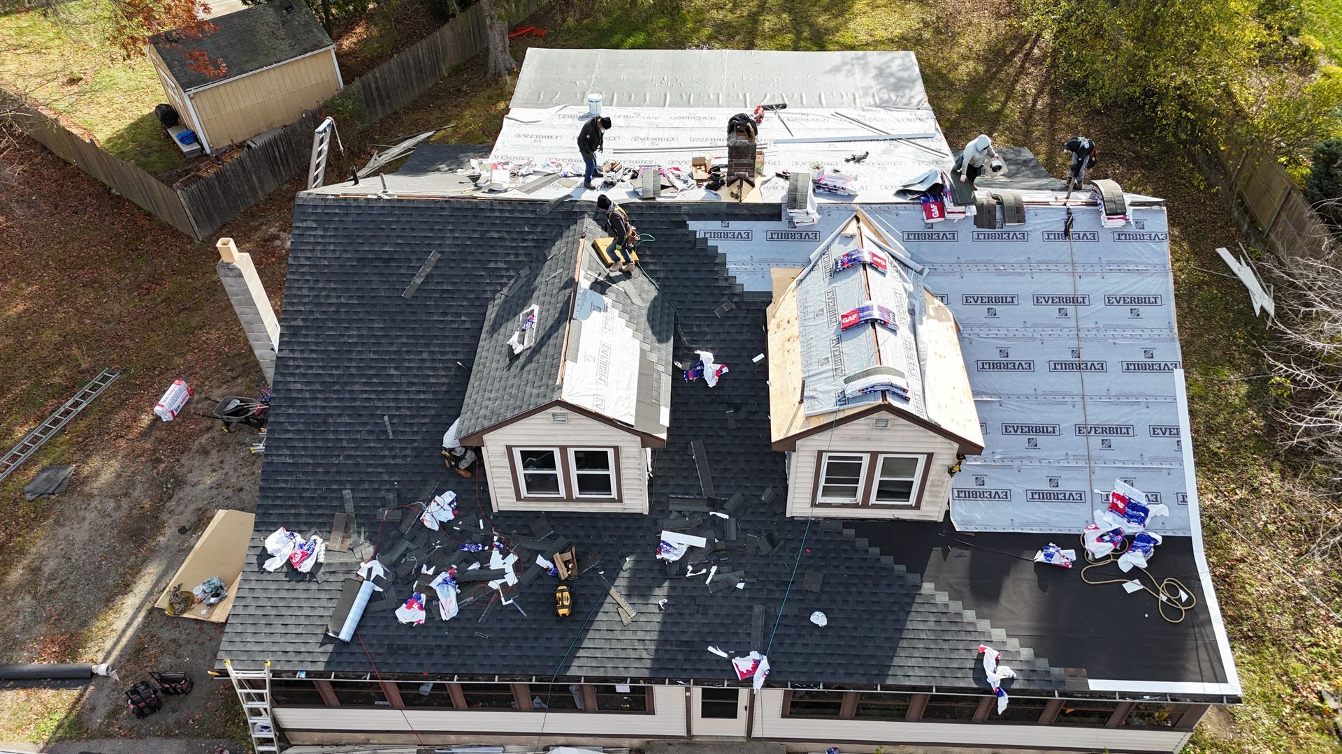 An aerial view shows multiple roofers working on a residential home's roof, replacing shingles and installing underlayment.