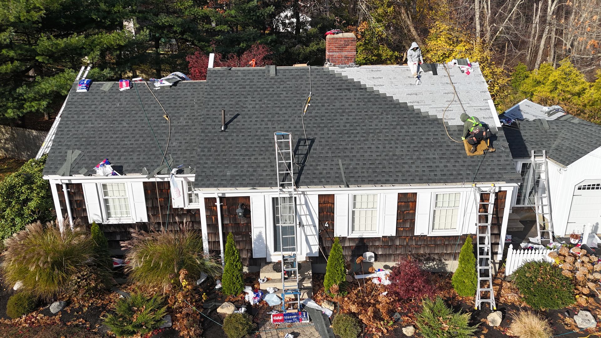 A high-angle view shows workers installing dark asphalt shingles on a residential house roof, leaving some area exposed.