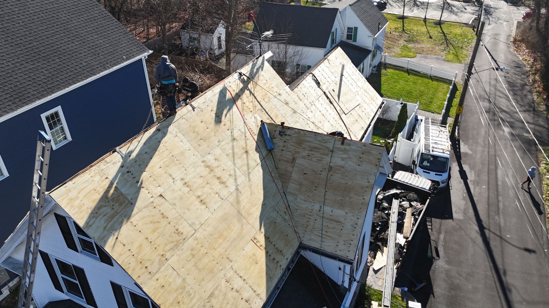 An aerial view shows workers installing new plywood roof decking on a home, with a construction van parked nearby.