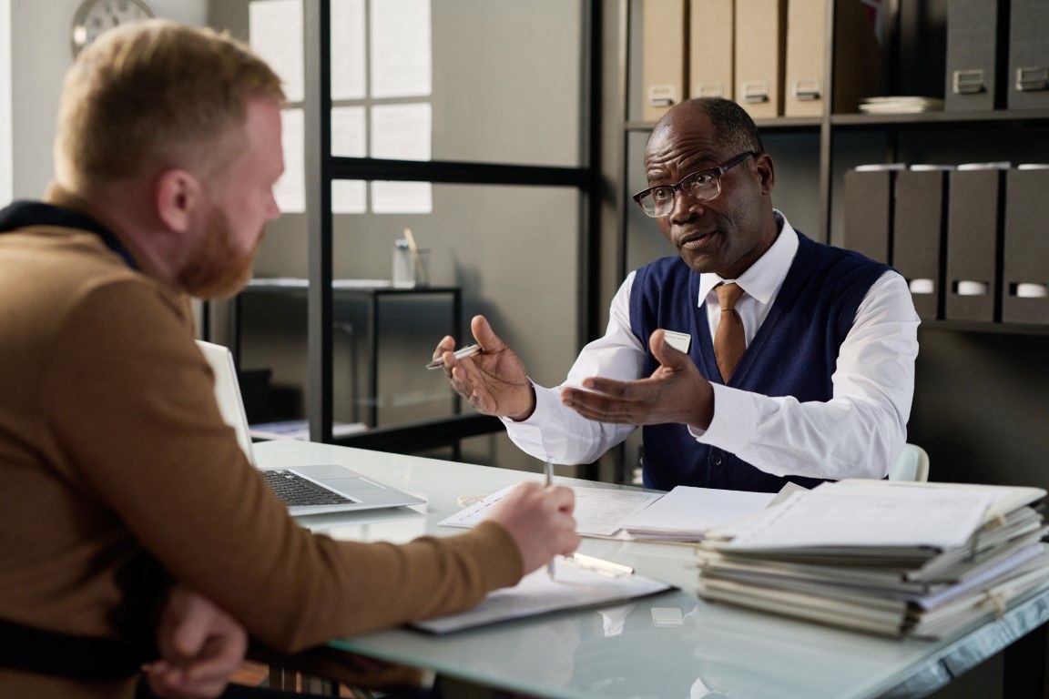 Two people in a meeting, one gesturing across a desk with papers in a modern office
