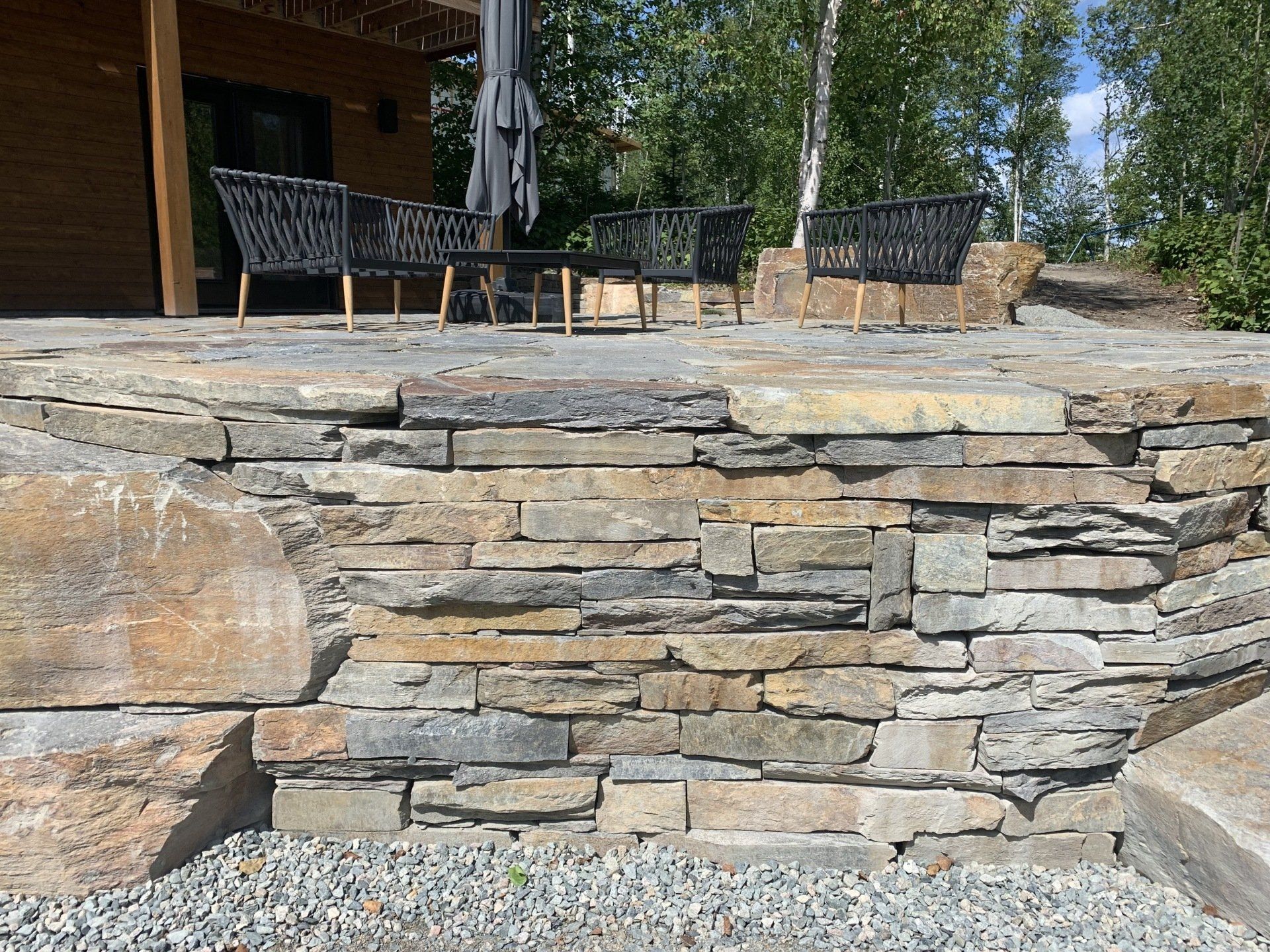 Mur de soutènement en pierre soutenant une terrasse avec mobilier de jardin, gravier gris en dessous.