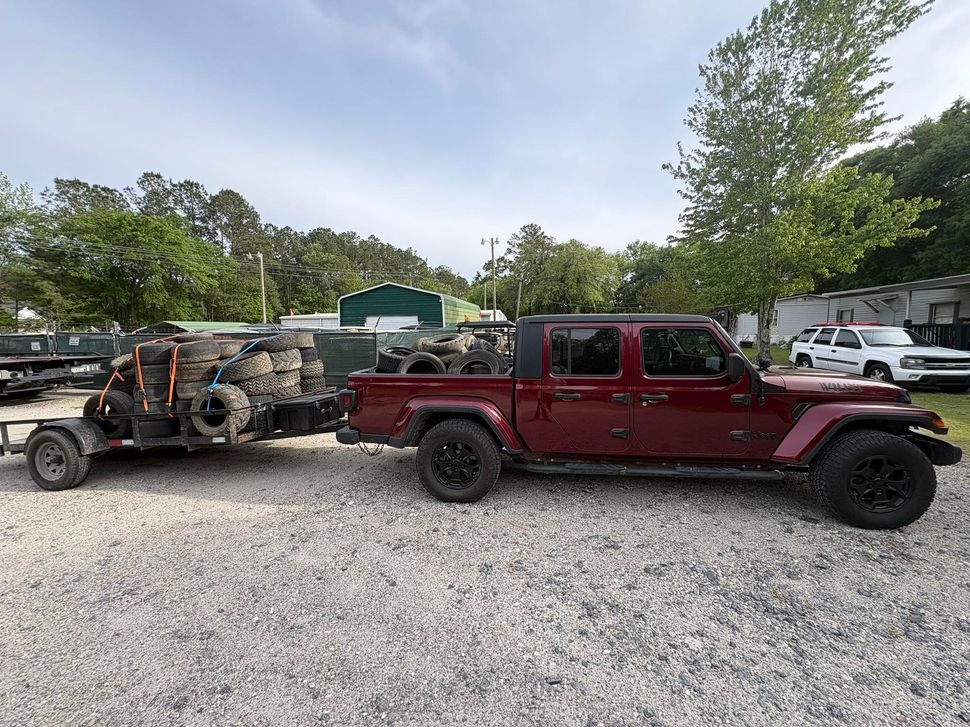 Maroon Jeep Gladiator towing a flatbed trailer in a gravel lot with trees and other vehicles nearby