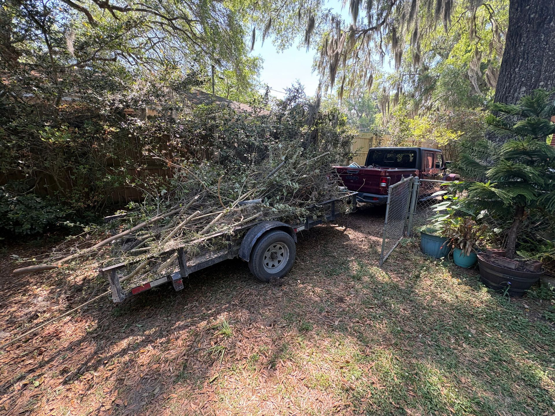 Flatbed trailer loaded with branches beside a red pickup truck in a wooded yard