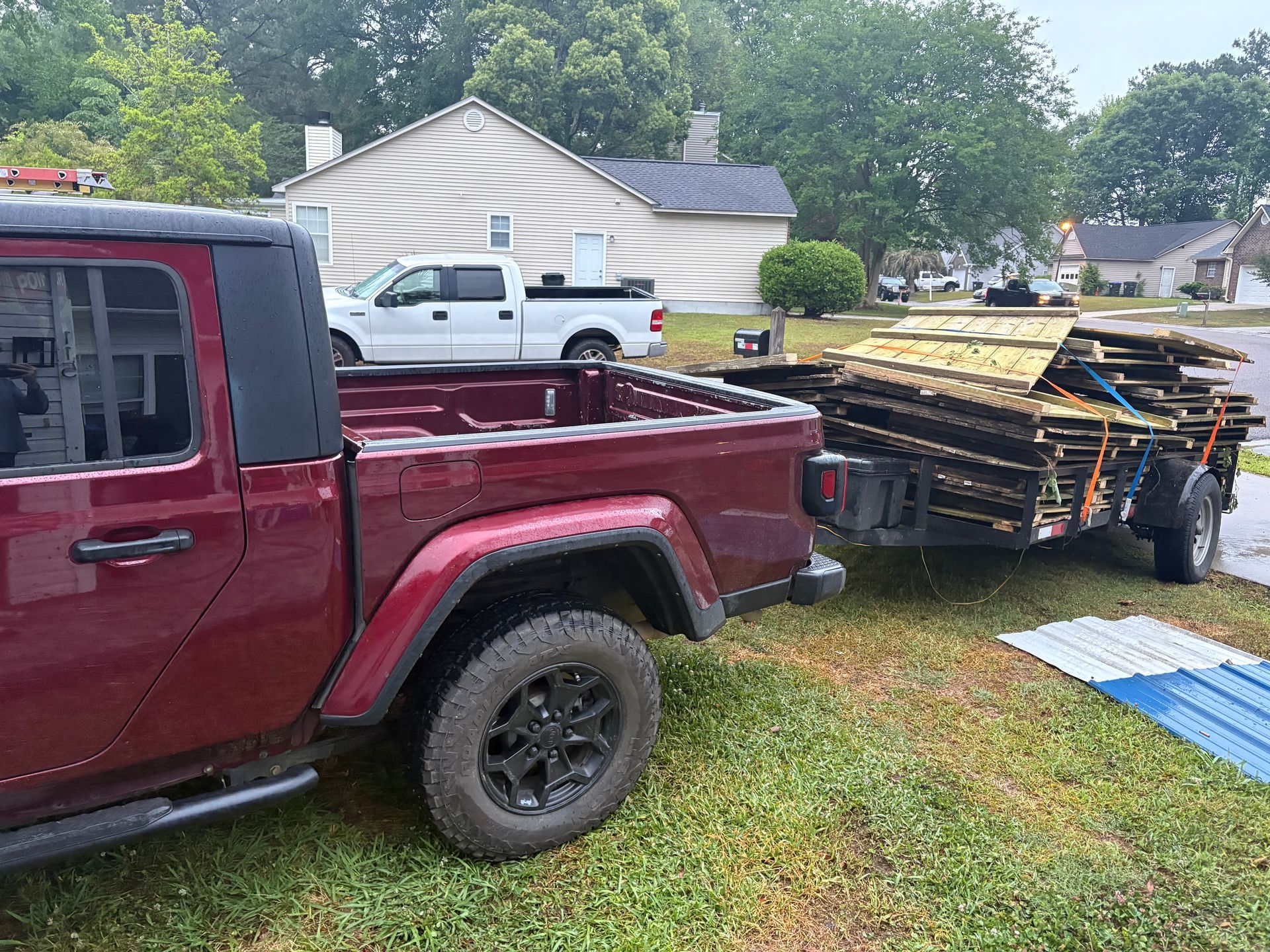 Red pickup truck towing a trailer loaded with stacked lumber on a grassy suburban lawn.