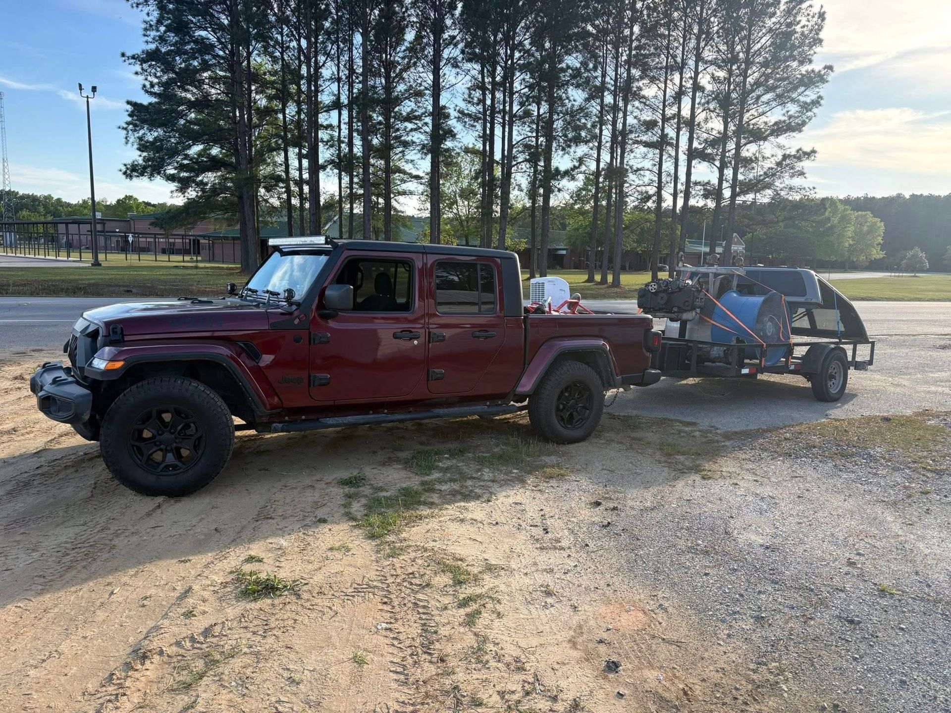 Red pickup truck towing a blue trailer on a gravel road near trees at sunset