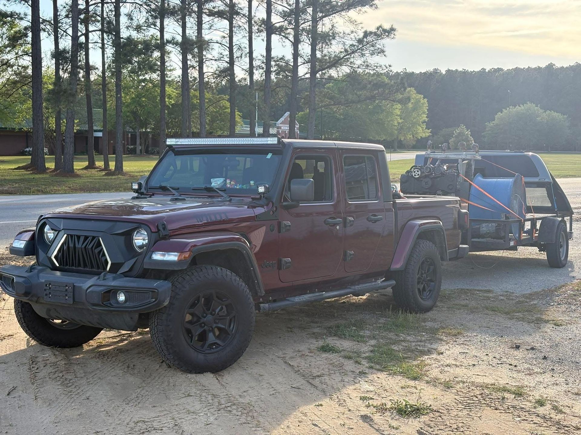 Maroon Jeep towing a blue off-road trailer on a dirt road in a wooded area.