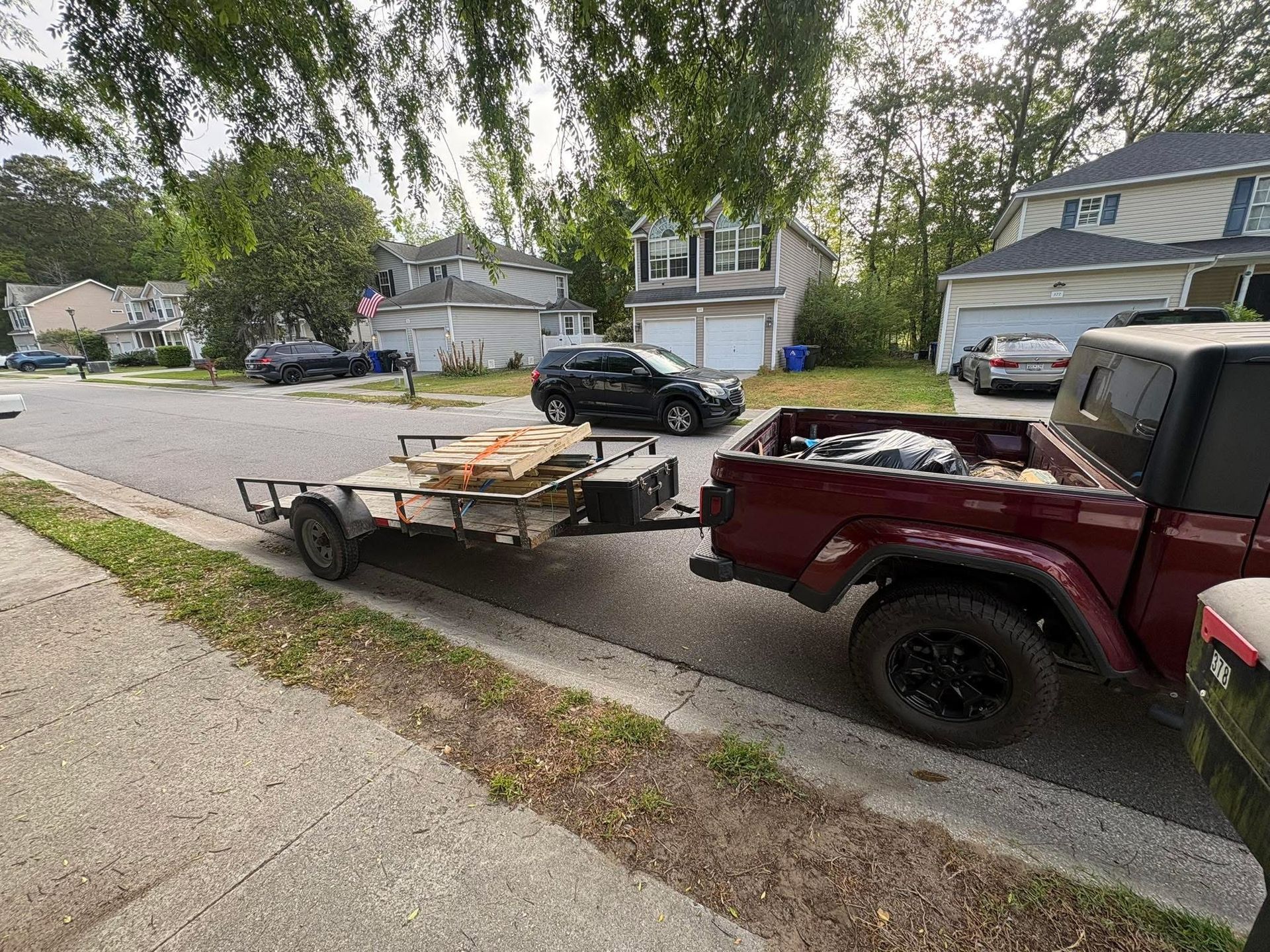 Red Jeep towing a small flatbed trailer along a suburban street lined with houses and trees