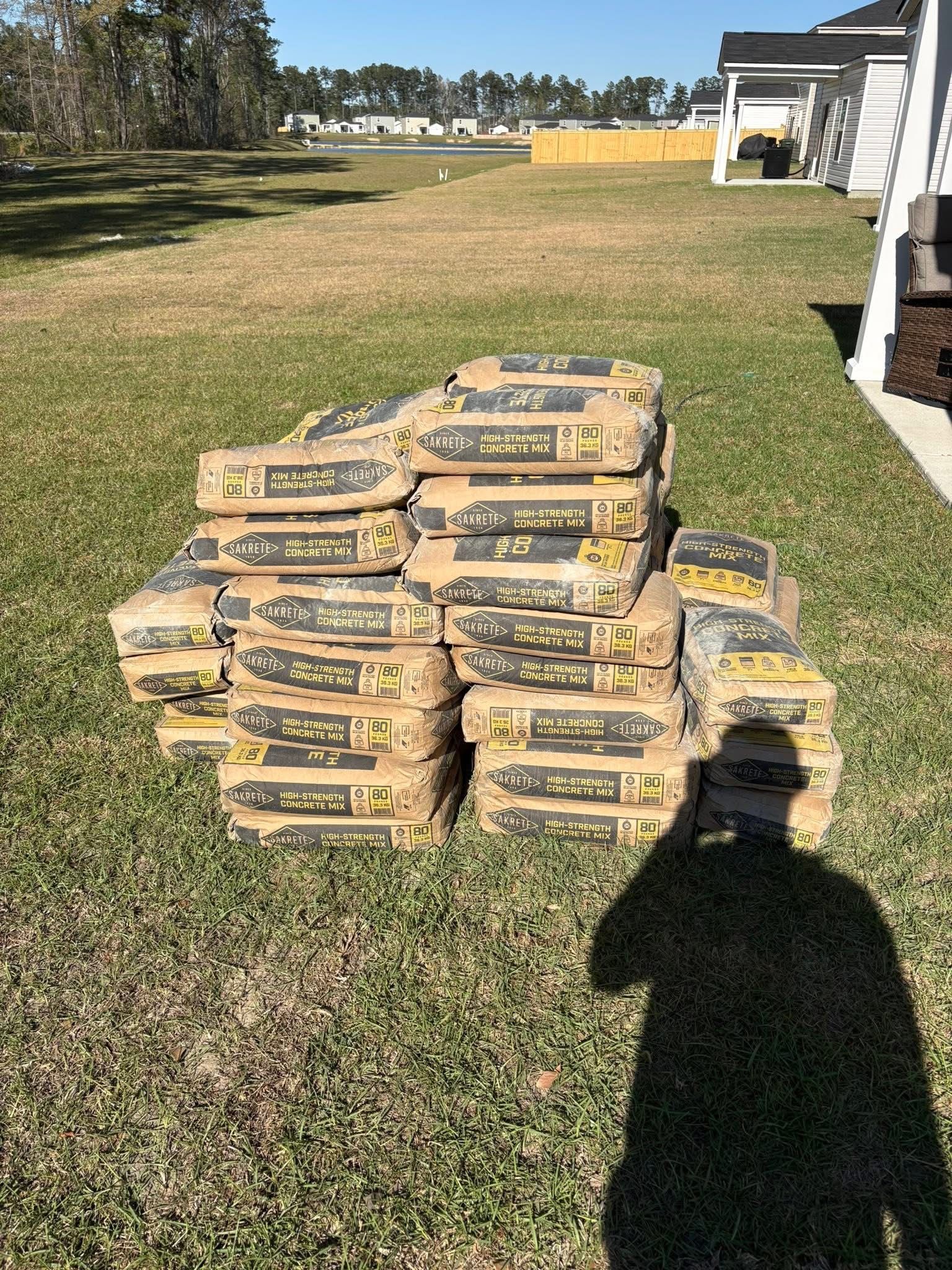 Stacks of bagged materials on a grassy lawn beside a house in bright sunlight.