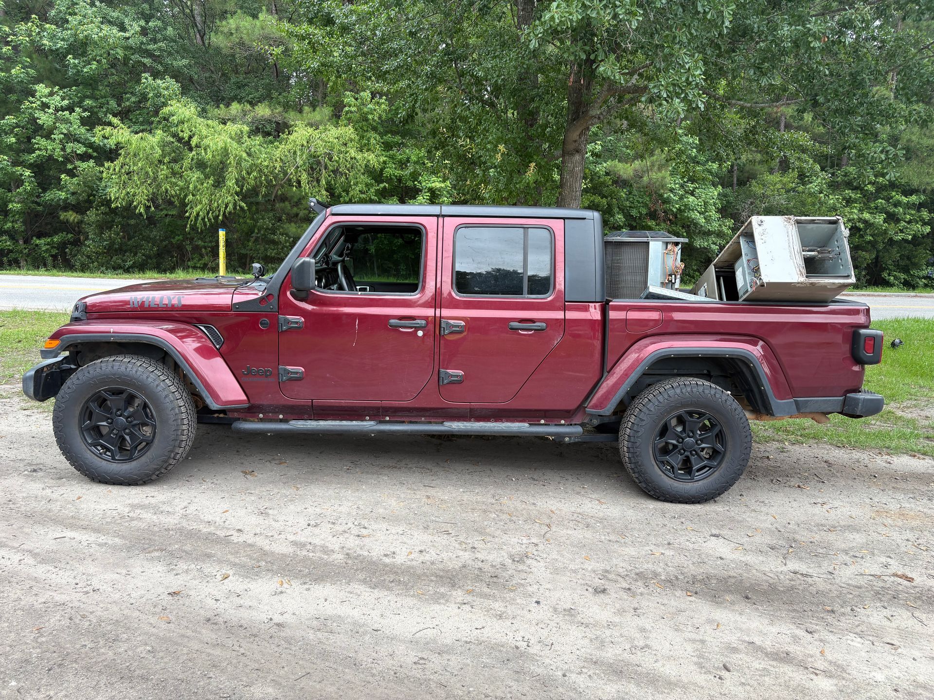 Maroon pickup truck parked on gravel with trees in the background, carrying items in the bed