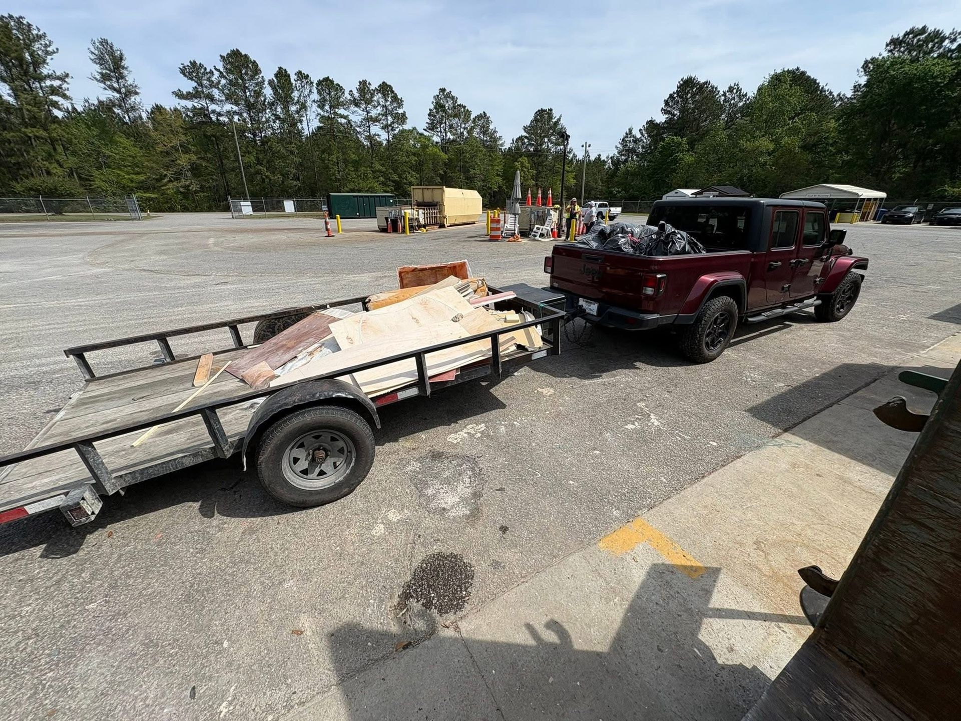 Maroon Jeep towing a flatbed trailer with debris in a gravel lot, trees and tents in the background