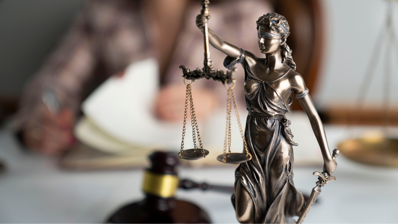 Lady Justice statue with scales in front of a blurred gavel and person writing in the background