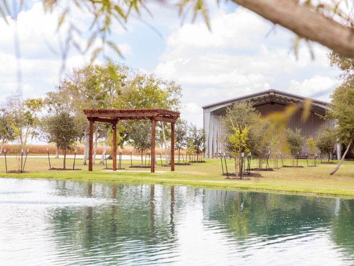 Wooden arbor beside a pond in a grassy field, with a metal building in the background.